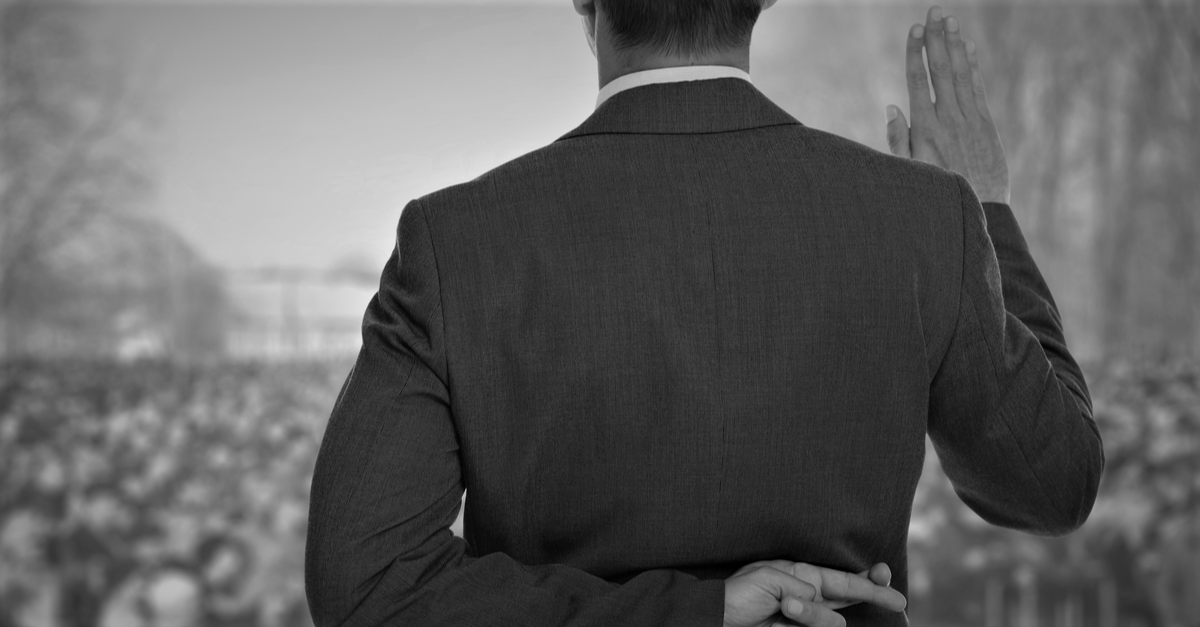 Man in suit with hand raised taking an oath in front of outdoor crowd, with other hand behind his back with fingers cross symbolizing contradiction and hypocrisy