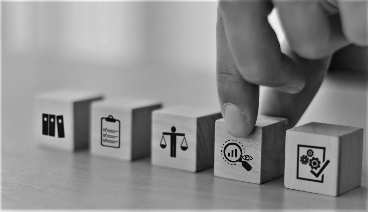 5 blocks on a table each with a different icon (from left to right): 3 books, clip-board to-do list, stick figure as a scale, magnifying glass, and a square with gears and a checkmark. Fingers go to pick up the block with the magnifying glass icon