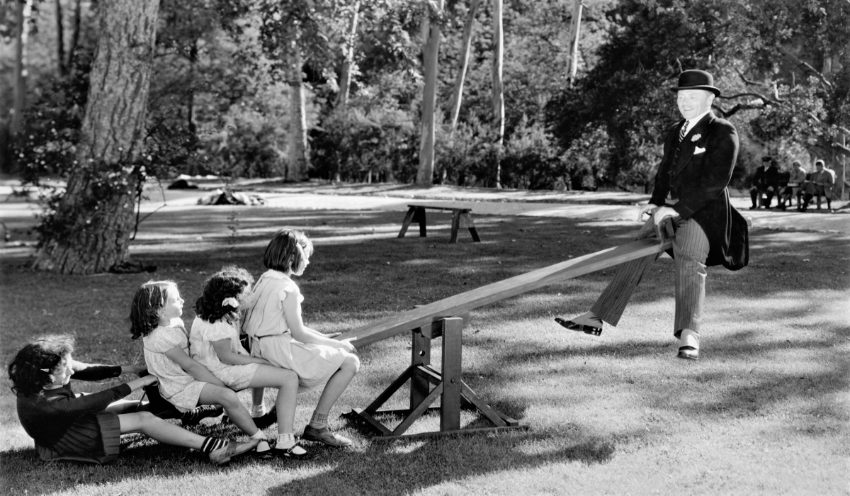 4 children on one end and a man on another end of a seesaw in a forrest-lined park (black and white)