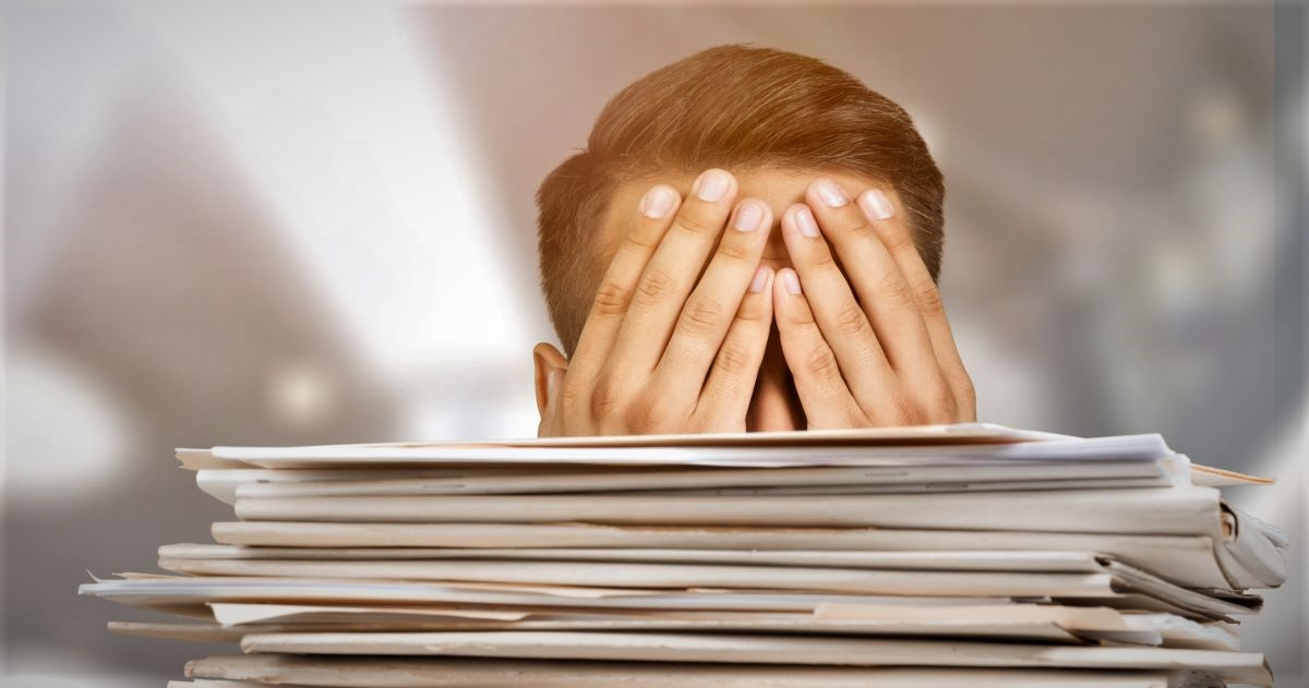 image of a man covering his eyes with a stack of paperwork in front of him